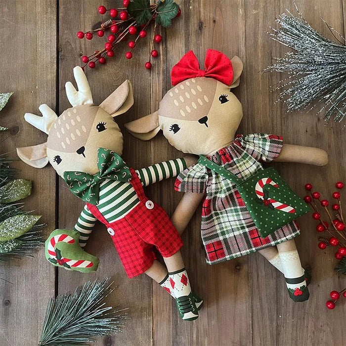 Two Christmas-themed plush reindeer dolls lying on a wooden surface, decorated with red berries, pine branches, and silver tinsel.