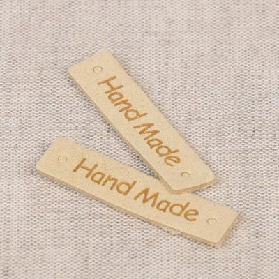 Two small rectangular light wood tags engraved with the words 'Hand Made' in brown, placed on a textured beige fabric background.