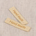 Two small rectangular light wood tags engraved with the words 'Hand Made' in brown, placed on a textured beige fabric background.