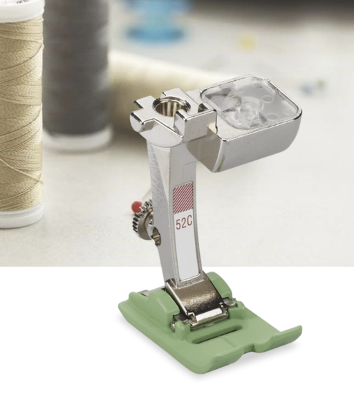 Bernina #52C Zigzag Presser Foot with a non-stick sole, placed on a sewing machine with spools of thread in the background.